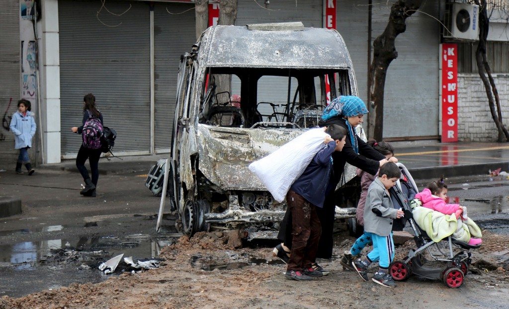 A woman with children walks by a vehicle, which was damaged during the clashes between security forces and Kurdish militants, in Baglar district, which is partially under curfew, in the Kurdish-dominated southeastern city of Diyarbakir, Turkey March 15, 2016. REUTERS/Sertac Kayar