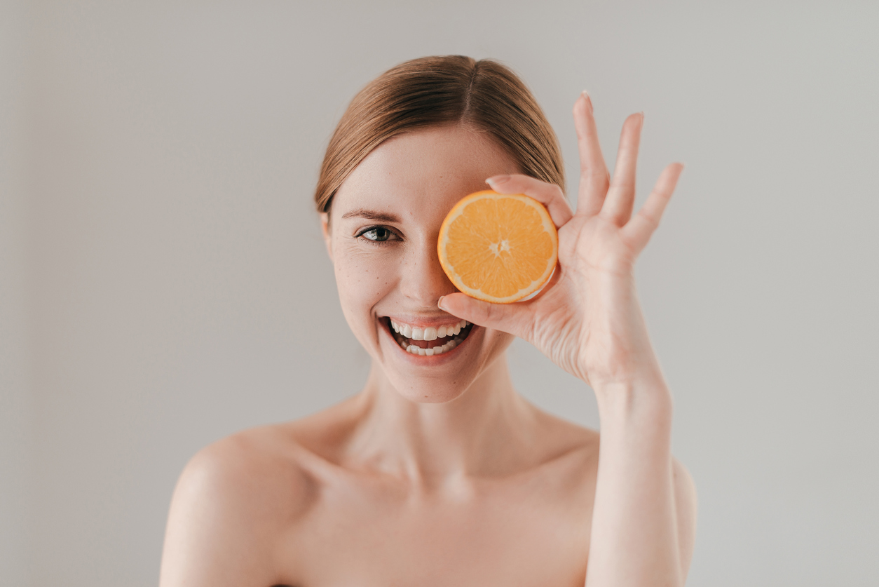Attractive young woman with freckles on face holding orange slice and smiling while standing against background