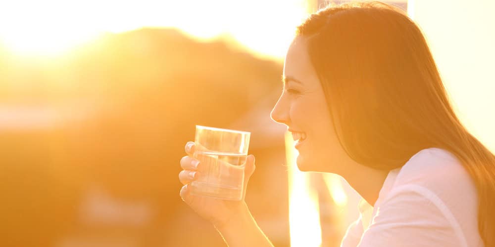 Side view portrait of a happy lady holding a glass of water outside in a balcony at sunset