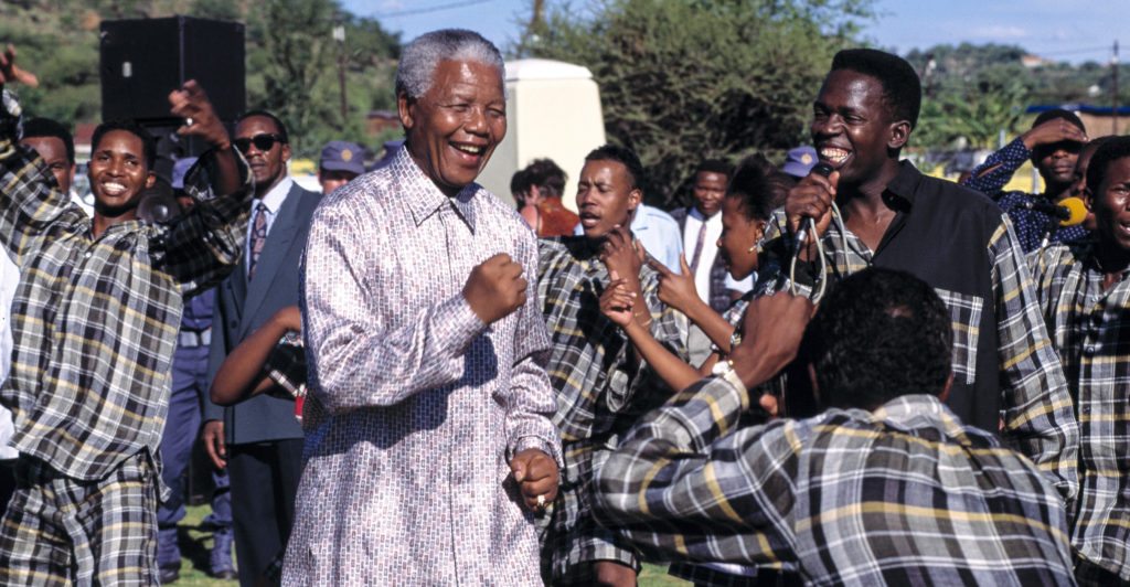 From Dare Not Linger, Youth welcome their hero as he demonstrates the famous ‘Madiba Shuffle’ while dancing to a local band during a visit to Oukasie township in Brits, 1995. Image copyright © Louise Gubb/lugubb@iafrica.com