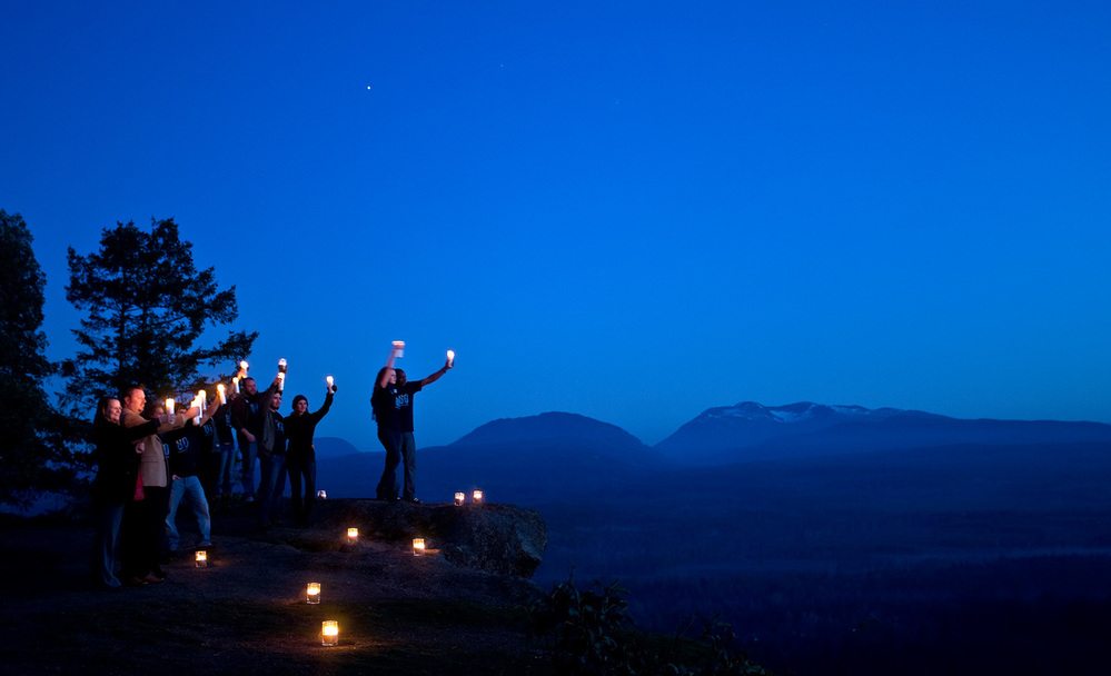 A group of men and women gathering on a clifftop on Vancouver Island against silhouetted trees and a twilight sky looking toward the sky and forested mountain scenery, Earth Hour 2010, Canada.