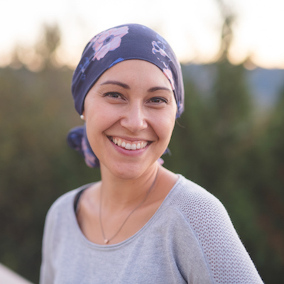A beautiful young ethnic woman wearing a head wrap looks toward the camera and smiles radiantly. She is standing outdoors and there are mountains and trees in the background.