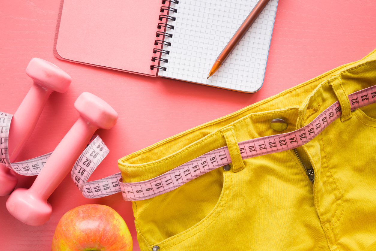 Measure tape with yellow jeans, dumbbells, apple, notebook on the pastel pink background. Women diet before summer season. Healthy lifestyle, body slimming, weight loss concept. Cares about body.