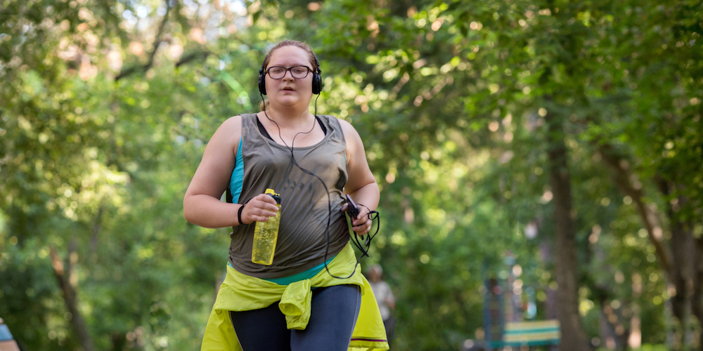Overweight woman running in the park with a bottle of water listening to music. Weight loss concept.