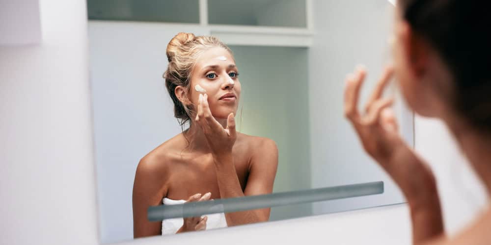 Young woman reflection in mirror applying cream in bathroom. Caucasian female doing beauty treatment on her face at home.