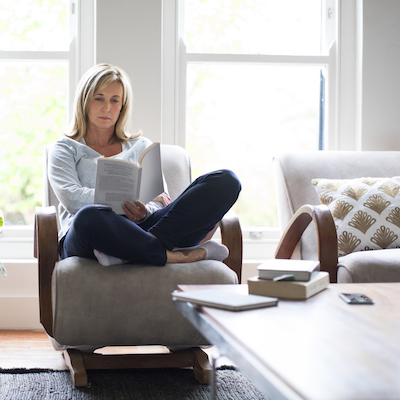 Full length of mature woman reading book on chair. Beautiful female is relaxing by houseplant against window. She is wearing casuals at home.