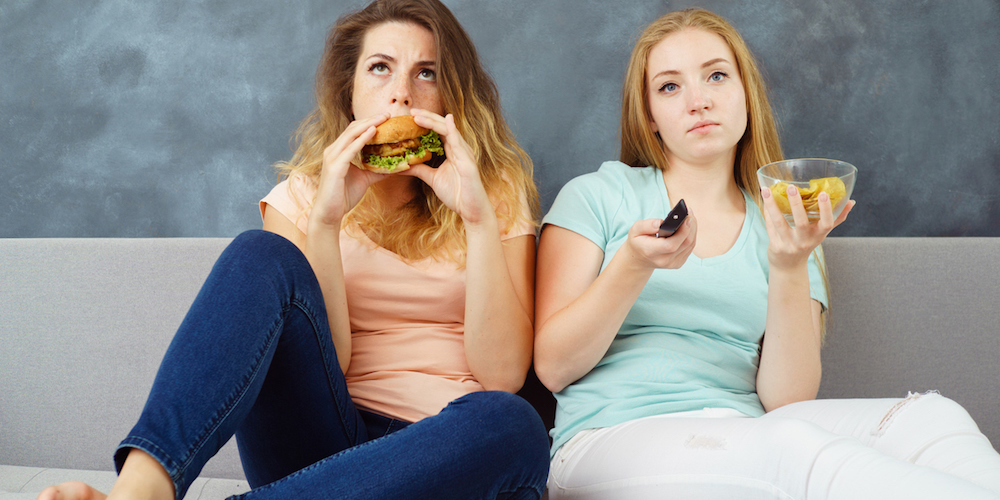 Overeating, mindless snacking, fast-food, party, delight, enjoyment, treat, appetite, hunger. Two cute young women sitting at coach with tv remote eating delicious burgers and chips greedily.