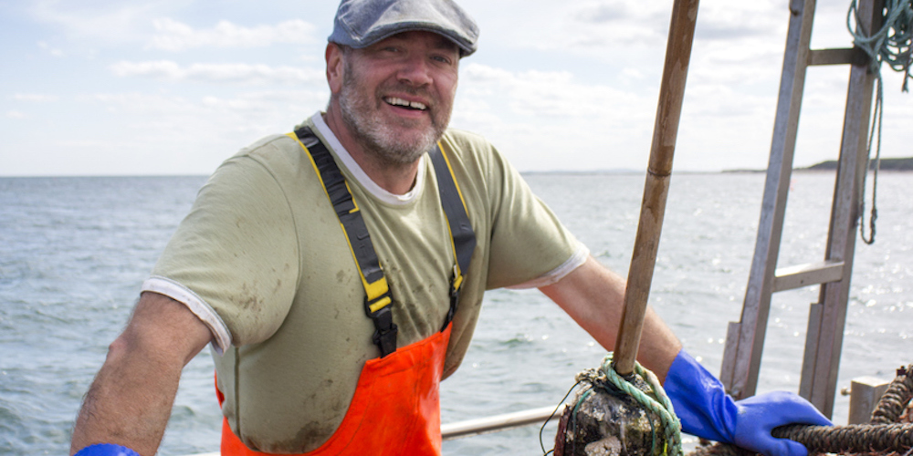 A happy fisherman smiles at the camera as he stands on his fishing boat. His lobster crate are propped up in front of him ready to be thrown into the sea.