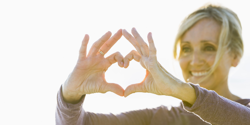 A mature woman in her 50s standing outdoors in the bright sunlight, holding her hands up in the shape of a heart to symbolize love and happiness. She is smiling. The focus Is on her hands.