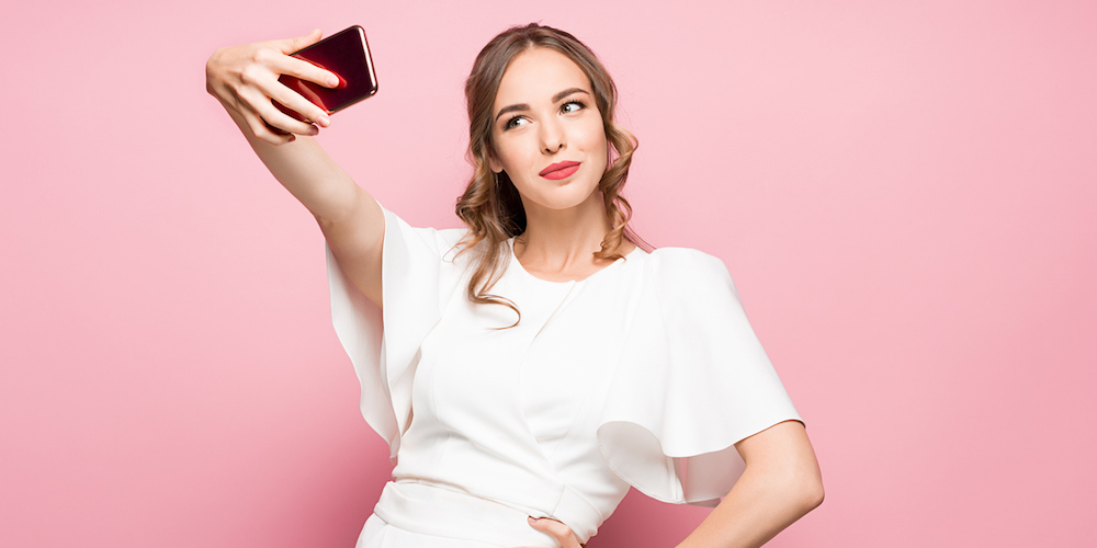 Portrait of a young attractive woman making selfie photo with smartphone on a pink studio background