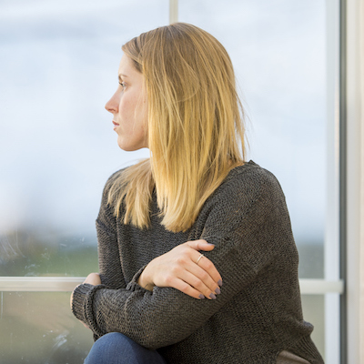 A caucasian woman in her 20s sits at the window and gazes outside. She has her arms crossed and is alone.