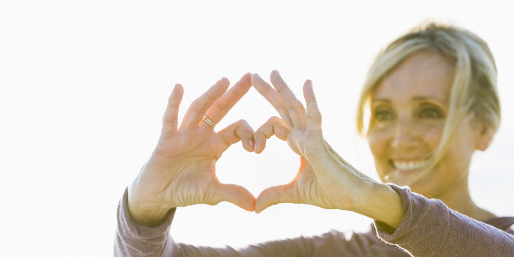 A mature woman in her 50s standing outdoors in the bright sunlight, holding her hands up in the shape of a heart to symbolize love and happiness. She is smiling. The focus Is on her hands.