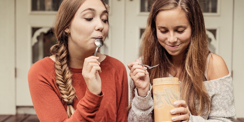 Two cute teenage girlfriends having fun eating peanut butter by the spoon on their house porch. Horizontal. Both girls have long light brown hair, one loose, the other with a braid. Both are wearing long sweaters in grey and orange. Waist up outdoors candid shot.