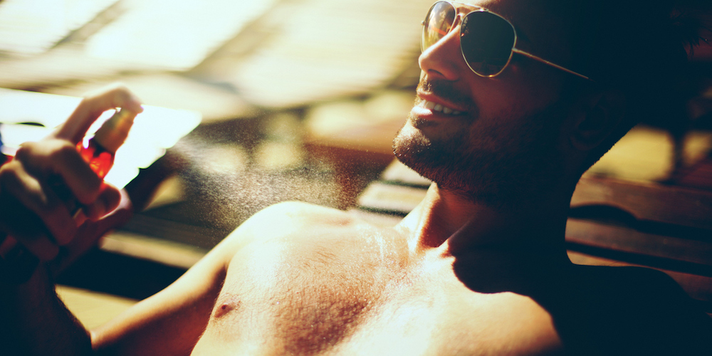 Handsome muscular guy relaxing out on a deckchair and using sunscreen.Applying it onto his body. He's wearing black swimsuit and sunglasses.He's smiling. Backlit.