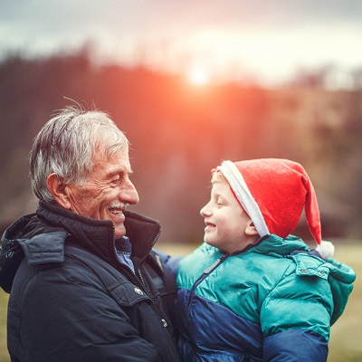 A portrait of a grandfather playing with his five year old grandson in the park. The old man is wearing a black jacket. The little boy is wearing a jacket and Christmas hat  They are smiling and having fun. In the background are trees and yellow leaves.
