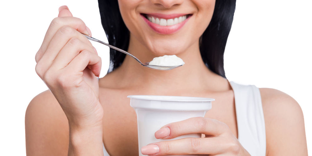 Close-up of young smiling woman holding a spoon with sour cream while standing against white background