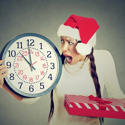 Closeup portrait worried stressed in a hurry young woman wearing red santa claus hat holding clock gift box isolated gray background. Emotion, funny face expression, last minute christmas shopping