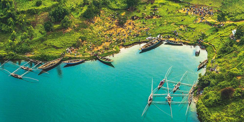 A large group of fishing boats is in display in Rwanda. They are docked at the edge of an island. A great number of people are seen gathering around the dock. Fishing rods can be seen hanging from the boats. Some small huts are visible around a set of trees. Getty Images