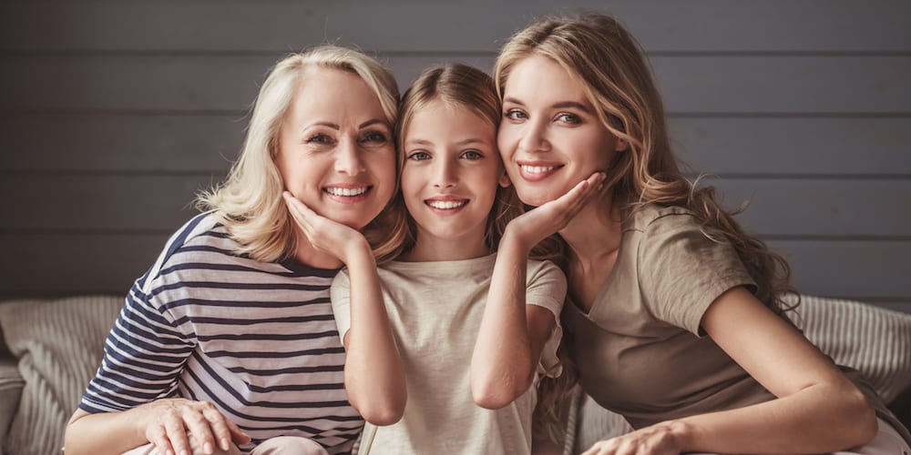 Beautiful women generation: granny, mom and daughter are hugging, looking at camera and smiling while sitting on couch at home (Beautiful women generation: granny, mom and daughter are hugging, looking at camera and smiling while sitting on couch at h