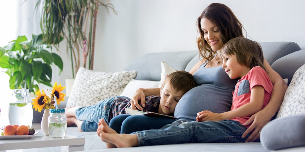 Young pregnant woman, reading a book at home to her two boys, eating fruits, hugging and laughing