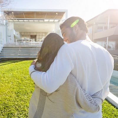 Young Couple standing in front of a modern new house. They are looking at the luxury home with their backs to the camera. They are sanding on a lawn and embracing.
