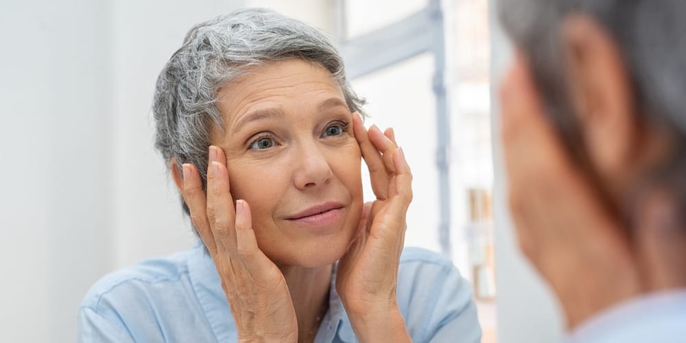 Beautiful senior woman checking her face skin and looking for blemishes. Portrait of mature woman massaging her face while checking wrinkled eyes in the mirror. Wrinkled lady with grey hair checking wrinkles around eyes, aging process. (Beautiful seni