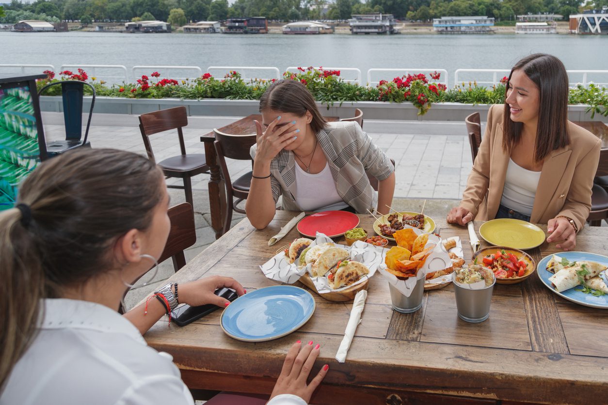 Young woman with hand over face looking embarrassed and laughing with friends, sitting in outdoor restaurant on city waterfront, Tex-Mex snacks on the table