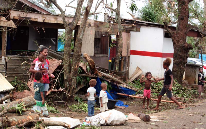 A woman carrying a baby stands with children outside homes damaged by Cyclone Pam, on a street surrounded by debris in Port Vila, the capital city of the Pacific island nation of Vanuatu