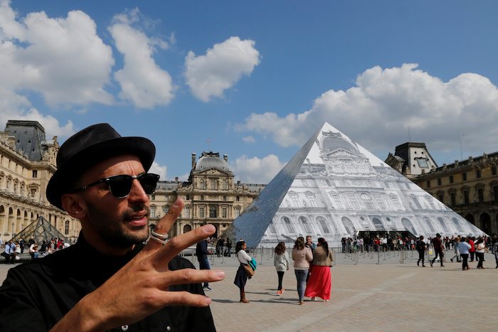 French artist JR stands in front of his lastest work, an image of the facade of Paris' Louvre covering the museum's pyramid entrance. May 25, 2016. REUTERS/Pascal Rossignol - RTSFWRP